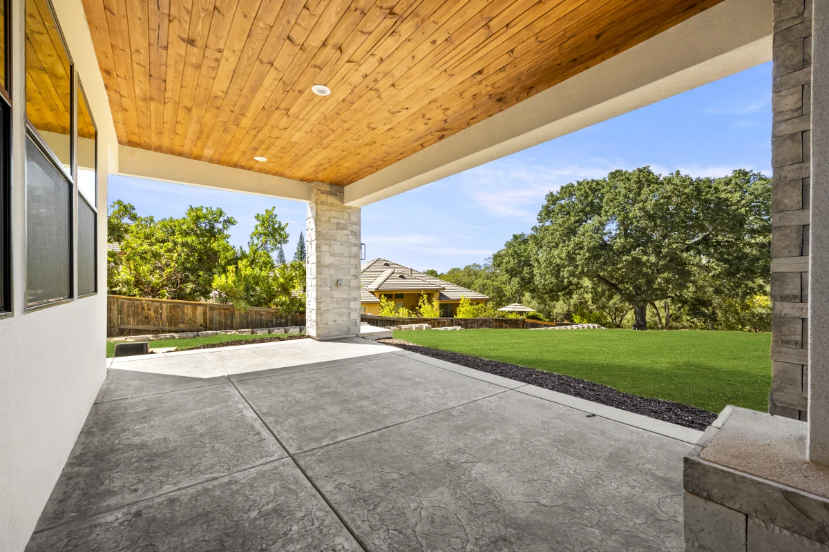 Under a solid patio cover with a cedar-plank ceiling and recessed lights, looking out across the lawn to the oaks