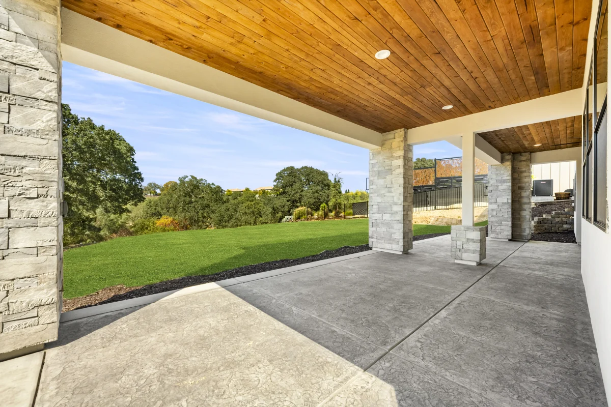Covered patio on a Solid-built home: a cedar-plank ceiling and stone columns over a stamped concrete slab, looking out to the lawn